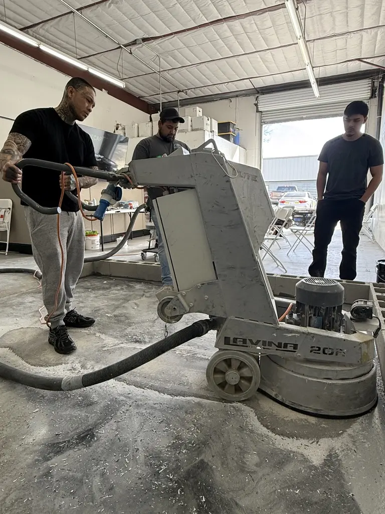 Two crew members running a walk-behind concrete grinder across a bare slab inside a warehouse