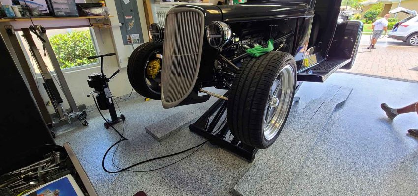 A black hot rod raised on a lift over a gray flake garage floor, tools along the wall and the driveway outside the open door
