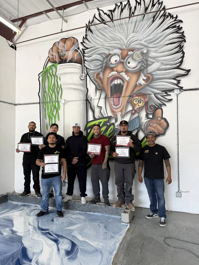 Eight people holding completion certificates in front of the shop mural, beside a freshly poured blue and white metallic test slab