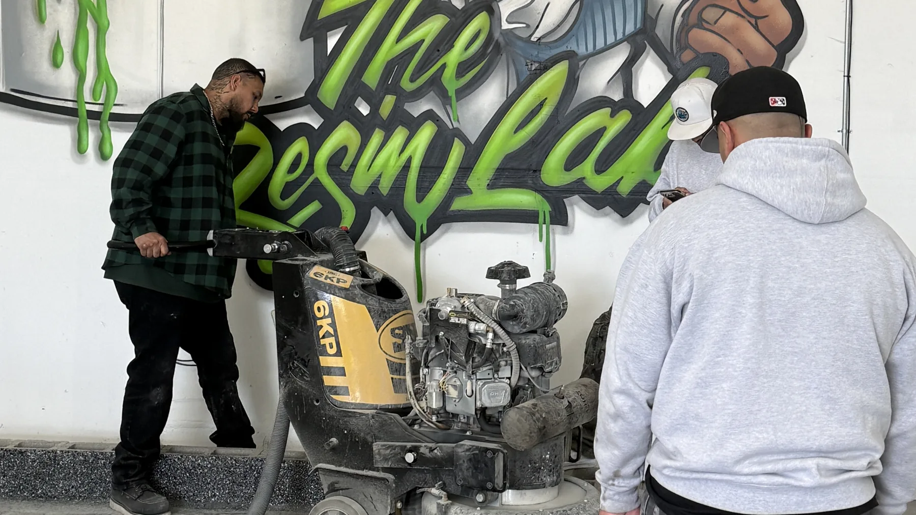 A crew member guiding a planetary grinder while two others watch, in front of a painted mural wall